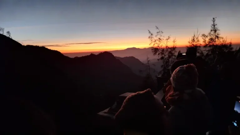 Pemandangan fajar subuh dengan siluet pegunungan dan langit gradasi jingga kekuningan dari atas penanjakan Gunung Bromo bersama para wisatawan.