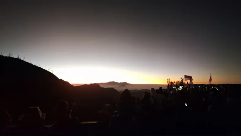 Pemandangan matahari terbit berwarna oranye di ufuk timur Gunung Bromo dengan siluet kerumunan wisatawan yang sedang berfoto di Penanjakan.